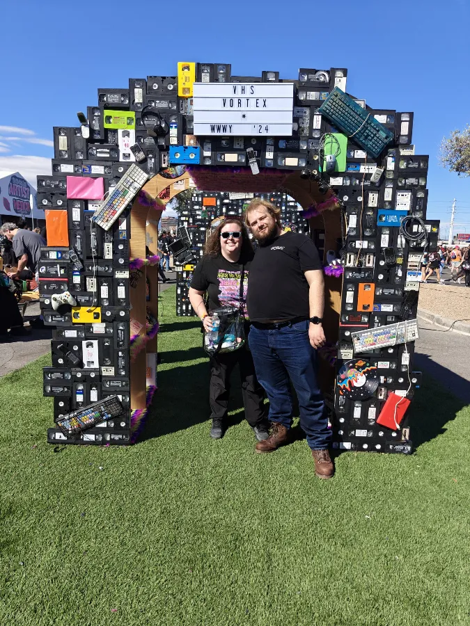 Michelle and her husband standing under an archway built from stacked VHS tapes and old electronics at an outdoor music festival.