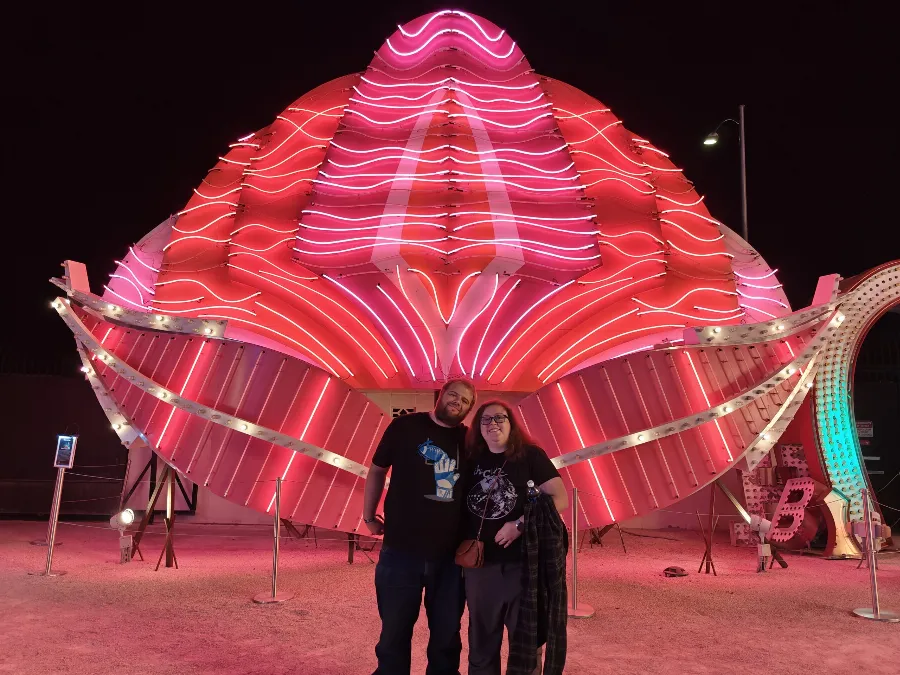 Michelle and her husband in front of a large pink and red neon sign at the Neon Museum in Las Vegas at night.